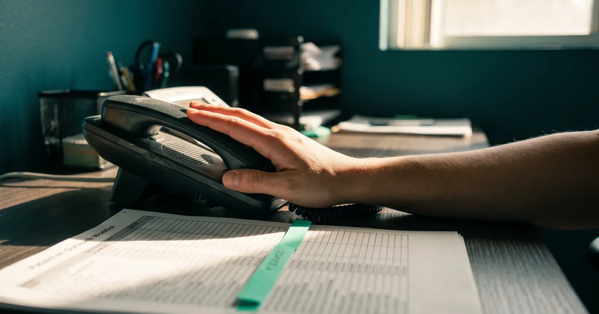 A hand resting on a desk phone handset beside a printed patient outreach worklist with a mint tab in a sunlit office